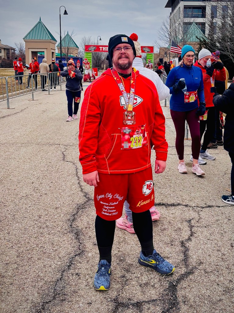 A man wearing a red Kansas City Chiefs sweatshirt and shorts, smiling at the camera after completing a 5K race. He has a medal around his neck and is surrounded by other runners and spectators in a cold, outdoor setting.